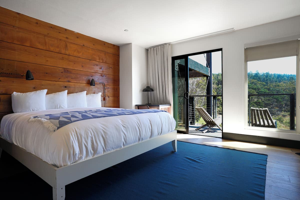 Quarto com uma cama grande, cabeceira de madeira, mesa de cabeceira, abajur e vista para o exterior através de uma porta de correr de vidro.