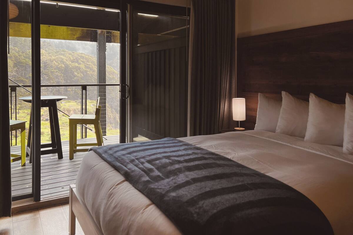 Quarto de hotel com cama, mesa de cabeceira, abajur, janela com vista para vegetação e porta deslizante para área externa.