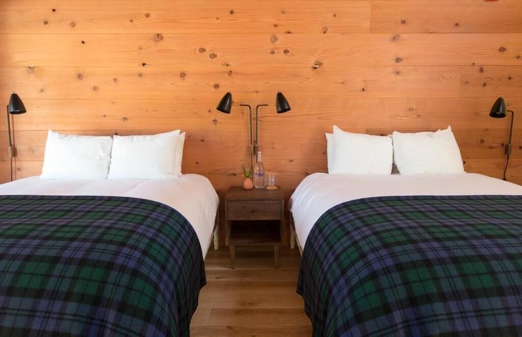 Une chambre double avec deux lits, chacun agrémenté de draps blancs et d’une couvre-lit à carreaux. Il y a deux tables de chevet avec des lampes, et un petit vase est posé sur l’une d’elles. La chambre est dotée d’une tête de lit en bois et d’un plancher en bois.