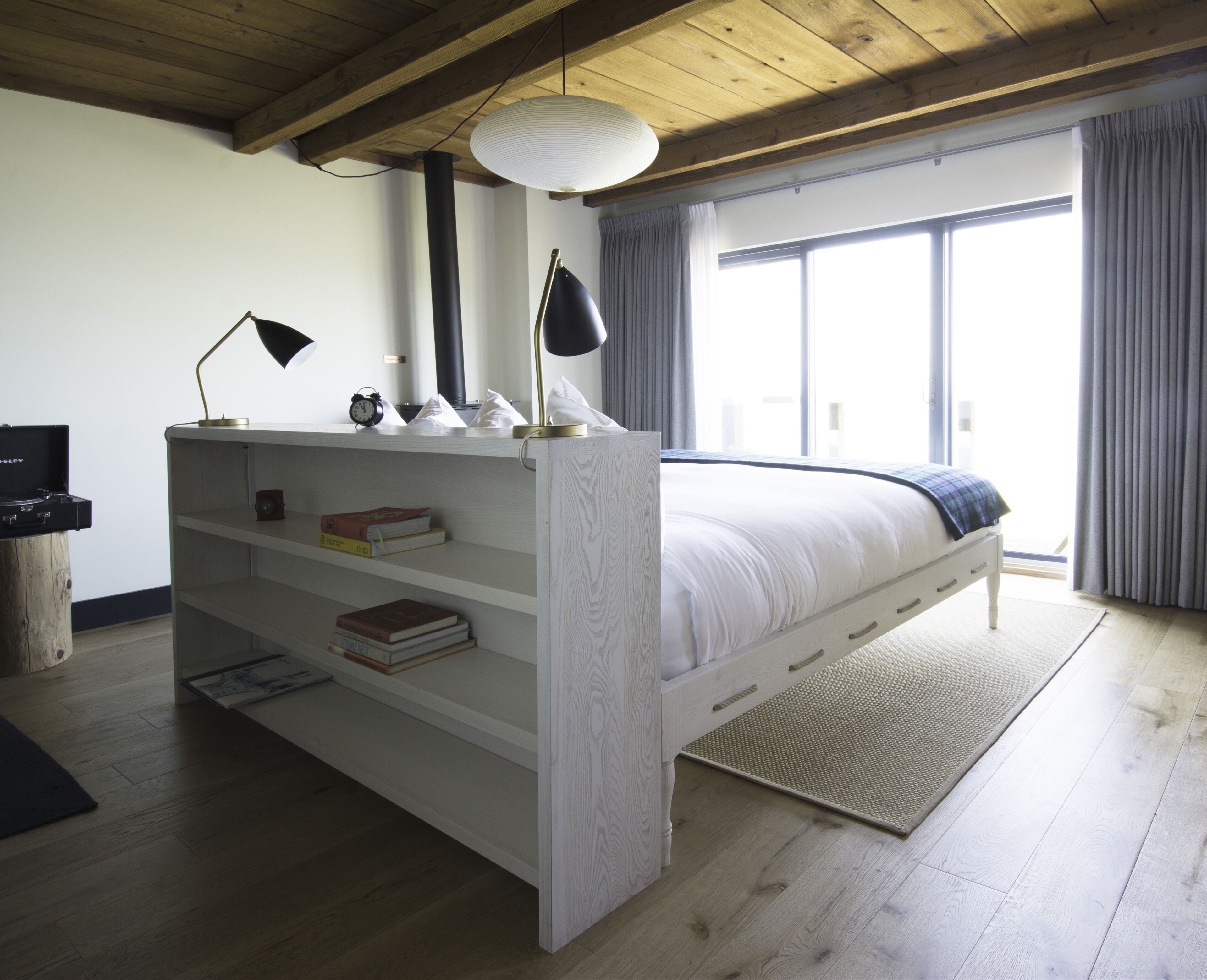A bedroom with a wooden ceiling, a bed with a white headboard, a bedside table with a lamp, and a window with curtains.