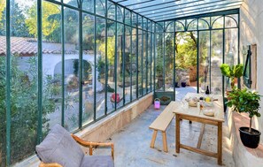 A conservatory with a glass roof, a wooden table, and chairs, surrounded by potted plants and a view of the garden. - Stunning home in Mornas (Mornas)
