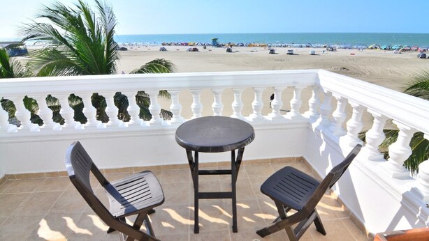 Varanda com duas cadeiras e uma mesa, com vista para uma praia com guarda-sóis e pessoas.