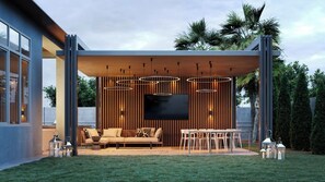 Un espacio exterior moderno con sofá, mesa de comedor y televisor, rodeado de listones de madera altos y luces colgantes.