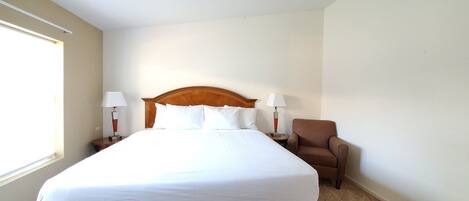 Quarto de hotel com uma cama grande, duas luminárias laterais, uma cadeira e uma janela.
