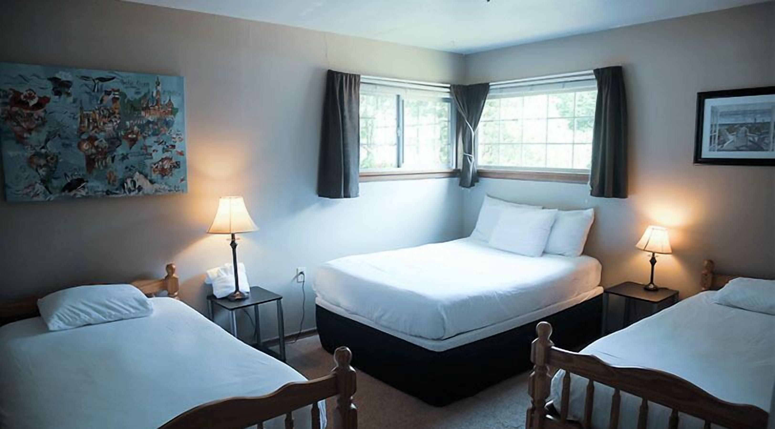 A hotel room with two single beds, a large bed, and a window with curtains. There is a painting on the wall and a lamp on each side of the large bed.