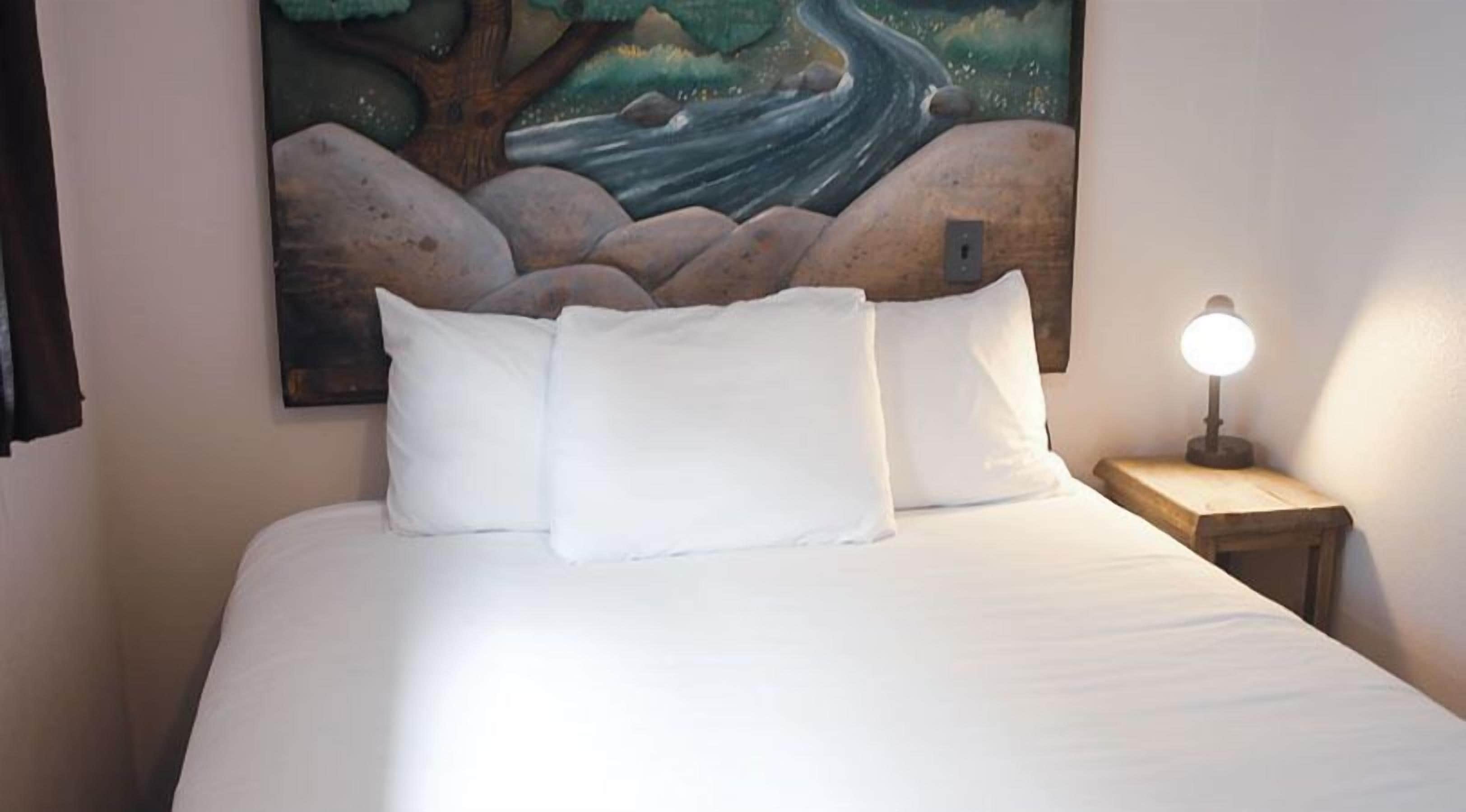 A bed with white linens and pillows, a nightstand with a lamp, and a painting of a river and rocks on the wall.