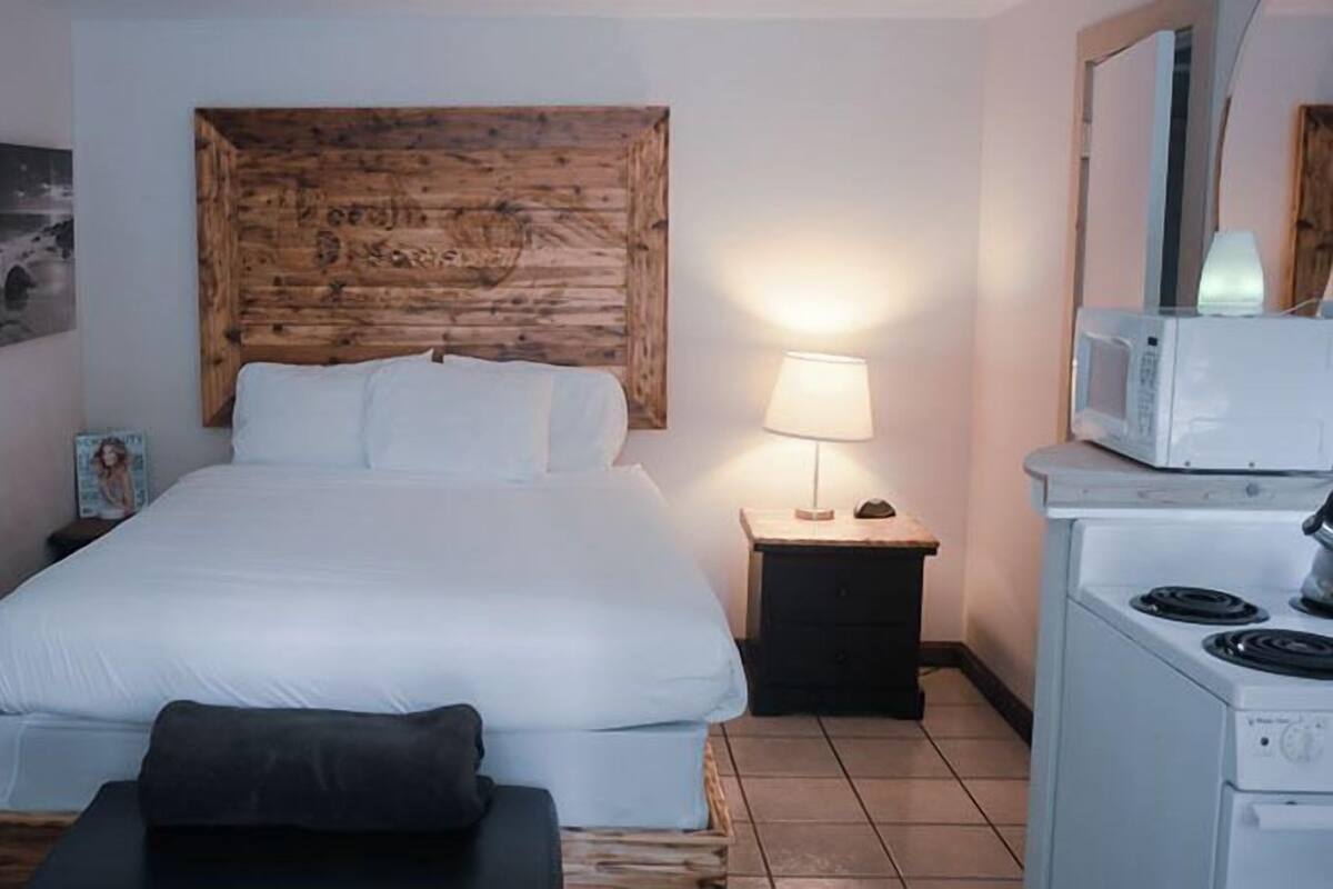 A hotel room with a wooden headboard, a bed with white linens, a nightstand with a lamp, a small table with a chair, and a microwave on a countertop.