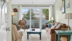 A cozy living room with a sofa, a coffee table, and a dining table. There is a sliding glass door leading to an outdoor area with a view of trees and a fence. - Quail Hollow A8-1U:  Beach Condo with Pool Access - Family Friendly (St. Augustine)