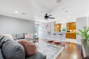 A modern living room with a grey sofa, a fluffy pink throw, and a coffee table. There are two bar stools and a ceiling fan. The kitchen in the background has wooden cabinets and a microwave.