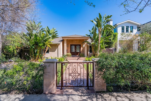 A single-story house with a front porch, surrounded by lush greenery and a gated entrance.