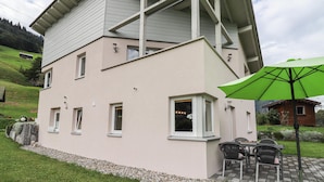 Ein zweistöckiges Haus mit geneigtem Dach, zahlreichen Fenstern und einer Terrasse mit Gartenmöbeln und einem grünen Sonnenschirm.