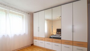 Ein Schlafzimmer mit großem Kleiderschrank, einem Bett und einem Fenster mit durchsichtigen Vorhängen.