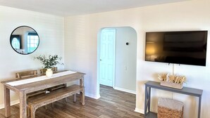 Een kamer met een houten bankje, een ronde spiegel, een aan de muur bevestigde televisie en een deur naar een andere kamer.