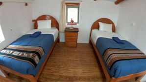 Ein Zimmer mit zwei hölzernen Betten, jeweils mit blauer Bettdecke und gemustertem Überwurf. Dazwischen steht ein kleiner Nachttisch aus Holz mit einer Lampe und einem Buch. Im Hintergrund ist ein Fenster mit Vorhang zu sehen.