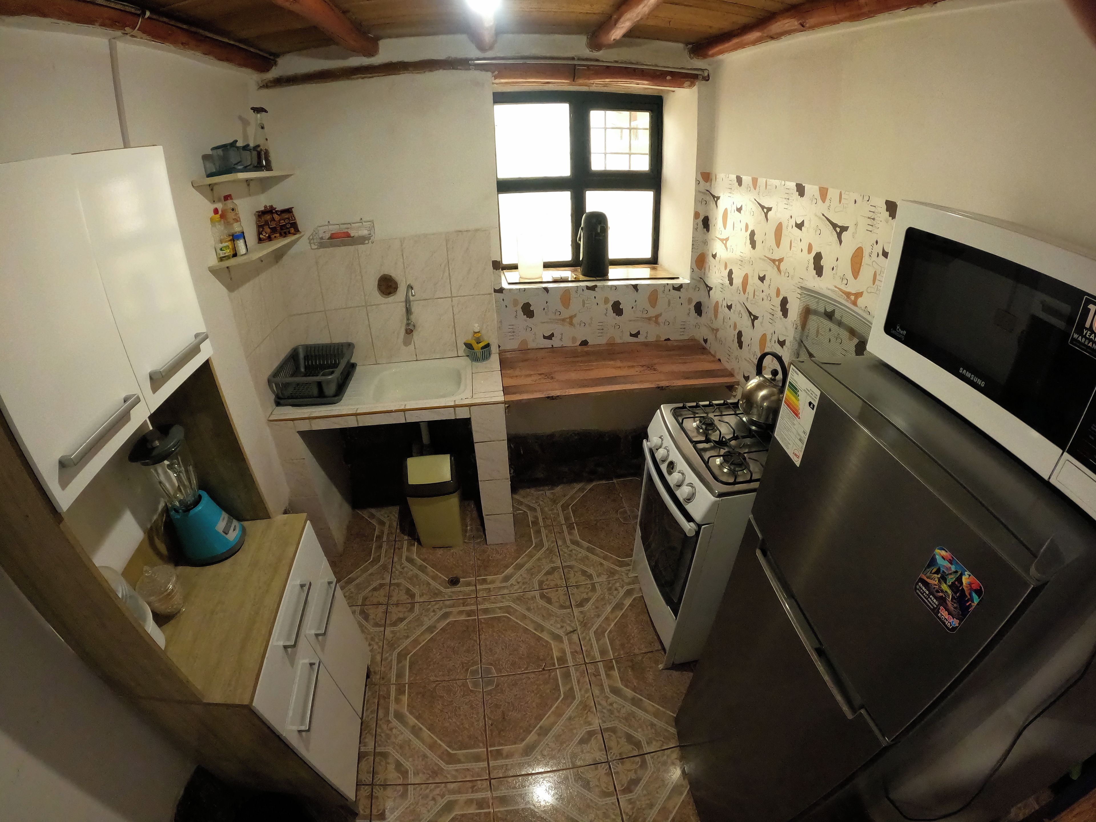 A compact kitchen with a gas stove, a microwave, and a refrigerator. There is a window above the sink, and the walls are tiled.