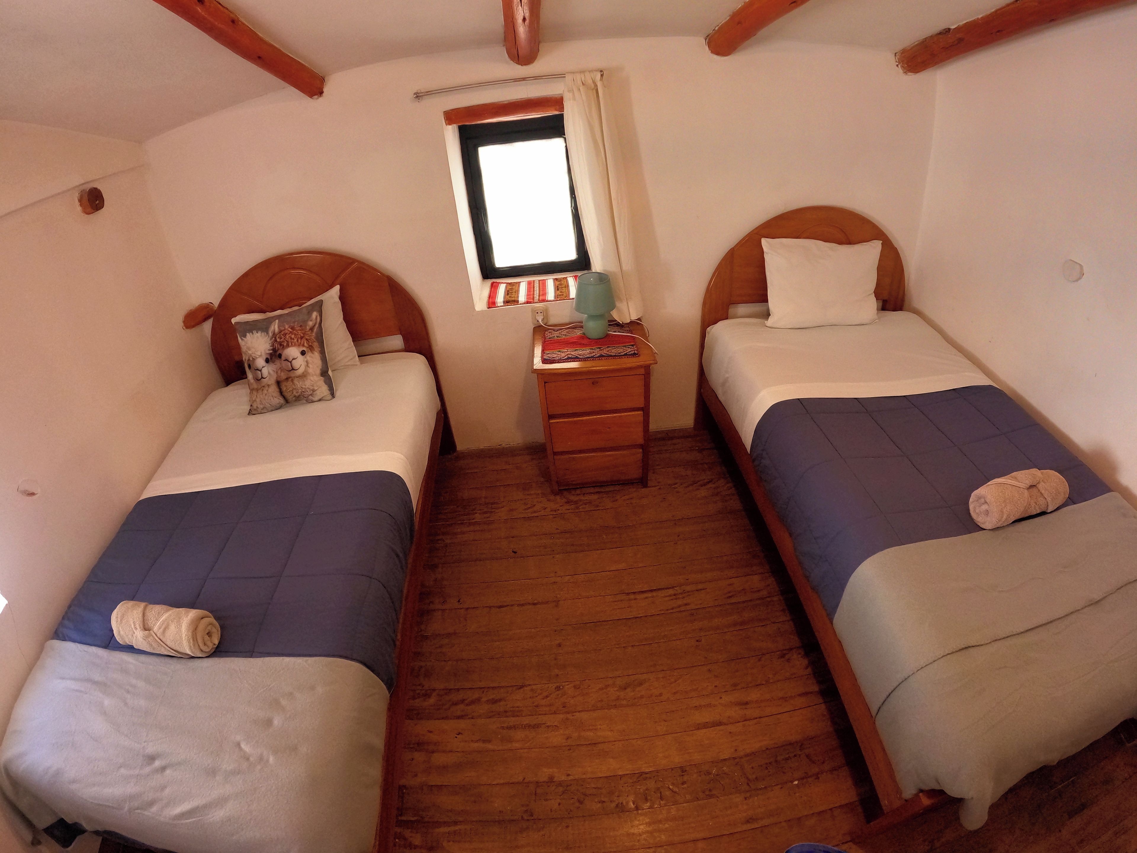 Une chambre avec deux lits simples, des têtes de lit en bois, une petite table de chevet avec une lampe et une fenêtre avec des rideaux.