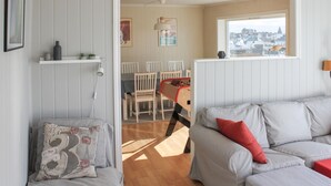 A living room with a grey sofa, a red pillow, and a white armchair. There is a window with a view of houses and a lamp on the wall. - 5 bedroom nice home in Borhaug (Borhaug)