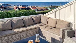 A sunlit outdoor seating area with a wicker sofa and table, overlooking a coastal town and the sea. - 5 bedroom nice home in Borhaug (Borhaug)