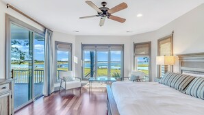 Un dormitorio amplio con una cama grande, un ventilador de techo y vistas al océano a través de ventanales de gran altura.