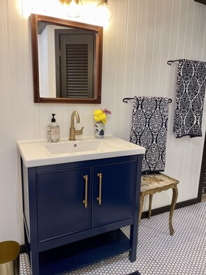 Un baño con tocador azul oscuro, lavamanos blanco, grifo dorado y un espejo encima. Hay un taburete pequeño con una toalla estampada y un cubo de basura al lado del tocador.