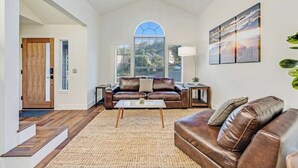 A spacious living room with a brown leather sofa, a coffee table, and a large window with a view of a cityscape. - Updated 5 BR house w/ hot tub - 5 min walk to downtown! (Geyserville)