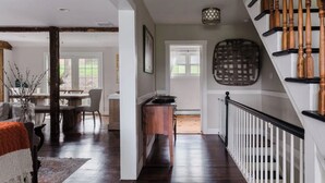Amplia sala de estar con piso de madera oscura, una mesa de comedor con sillas, un sofá y una mesa de centro. Se observa una escalera con balaustres de madera y barandilla blanca.