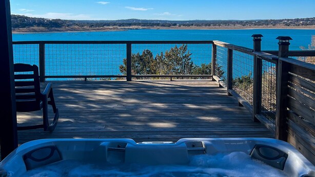 A wooden deck with a hot tub, overlooking a clear blue lake and distant hills.