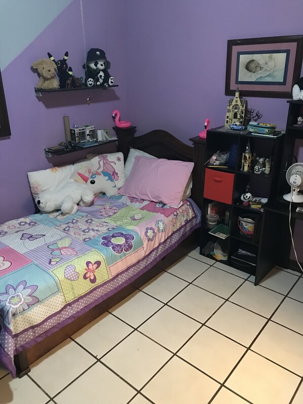 A bedroom with a bed, pillows, a quilt, a shelf with toys, and a fan.