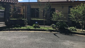 Un edificio amurallado con una cerca metálica, un camino pavimentado al frente y algo de vegetación.