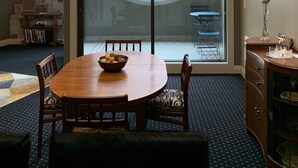 A dining area with a round table, chairs, a bowl of fruit, and a view of a brick building through a large window. - Henry Matthews Apartment (Dunedin)