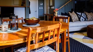 A dining area with a wooden table, chairs, a bowl of fruit, a teapot, and a mug. There is a staircase in the background leading to another room. - Henry Matthews Apartment (Dunedin)
