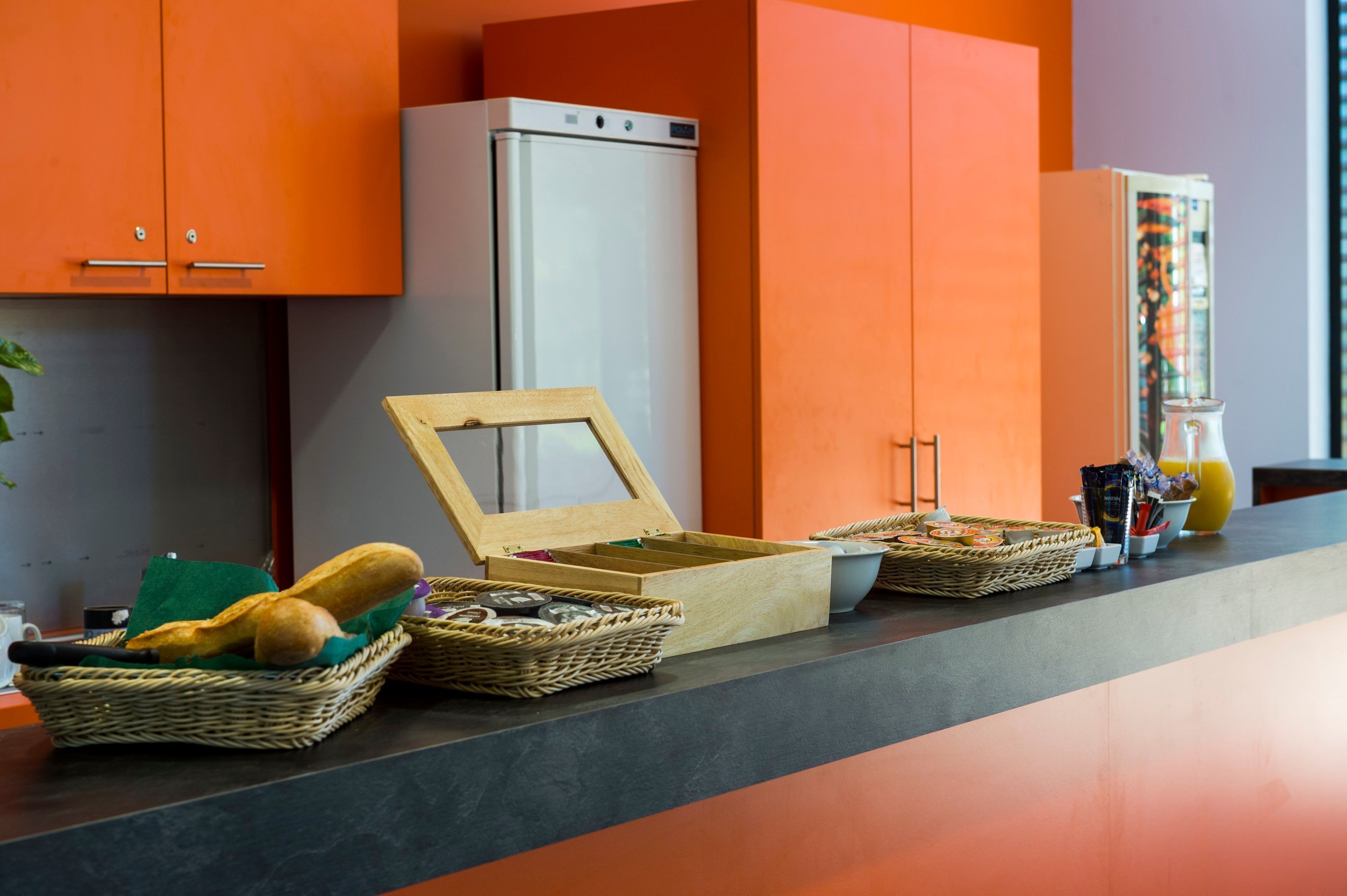 Eine Küche mit orangefarbenen Schränken, einem weißen Kühlschrank und verschiedenen Körben mit Brot und anderen Gegenständen auf der Arbeitsfläche.