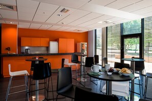 Une cuisine moderne avec des meubles de couleur orange et un plan de travail noir. On y trouve plusieurs chaises noires et une chaise blanche disposées autour d’une table sur laquelle se trouvent une tasse blanche et un sandwich.