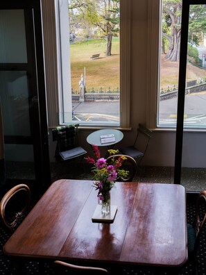 A dining area with a wooden table and chairs, a vase with flowers, and a view of a park through large windows. - John Campbell Apartment (Dunedin)