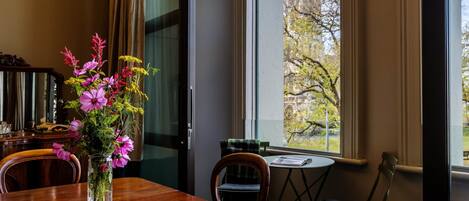 A dining area with a wooden table, chairs, a vase of flowers, and a view of trees and a building through a window.