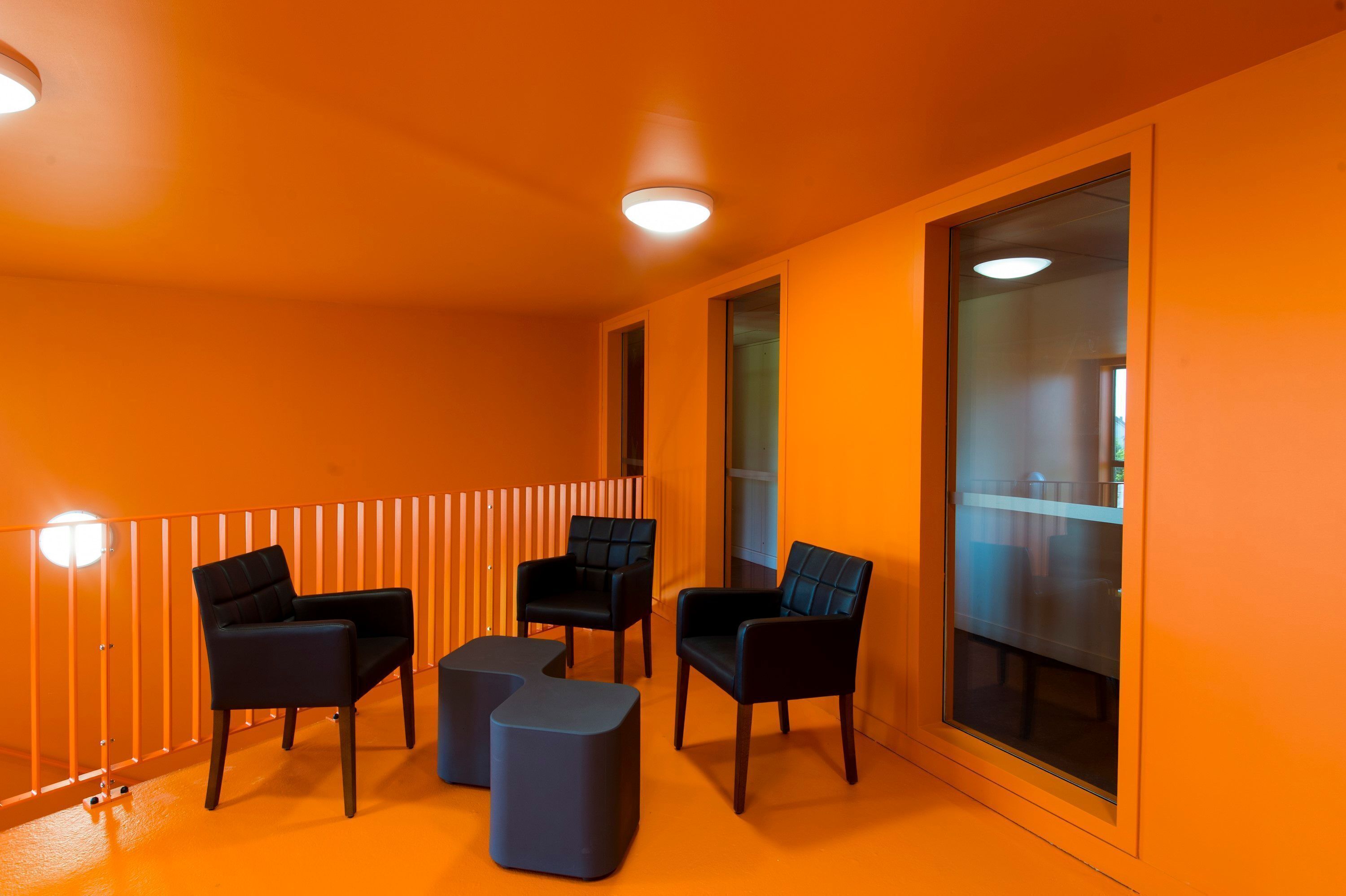 Een kamer met oranje wanden, een glazen scheidingswand en drie zwarte stoelen met een klein voetenbankje.