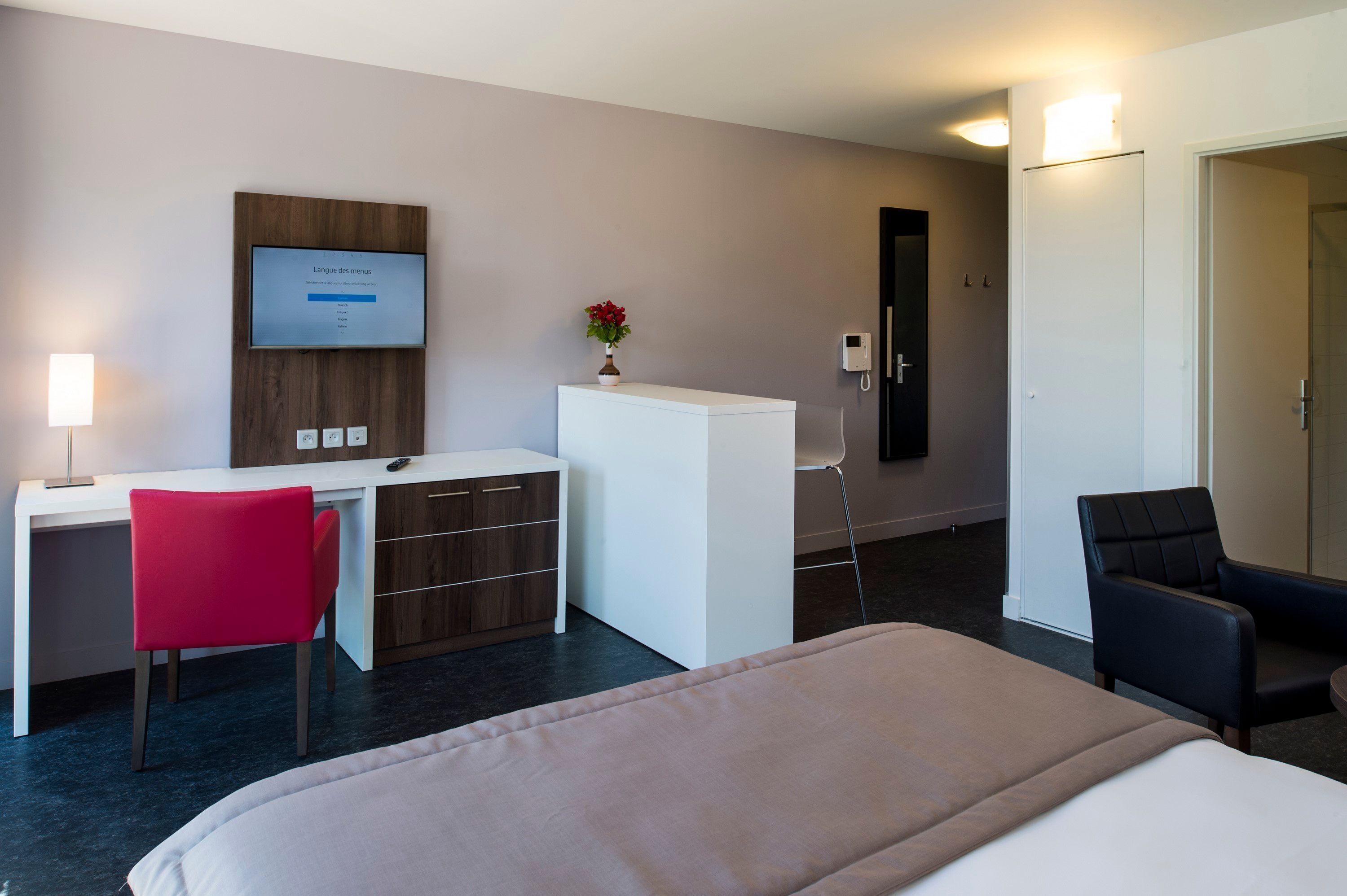 A hotel room with a bed, a desk with a chair, a small white cabinet, a TV mounted on the wall, a vase with flowers, and a door.