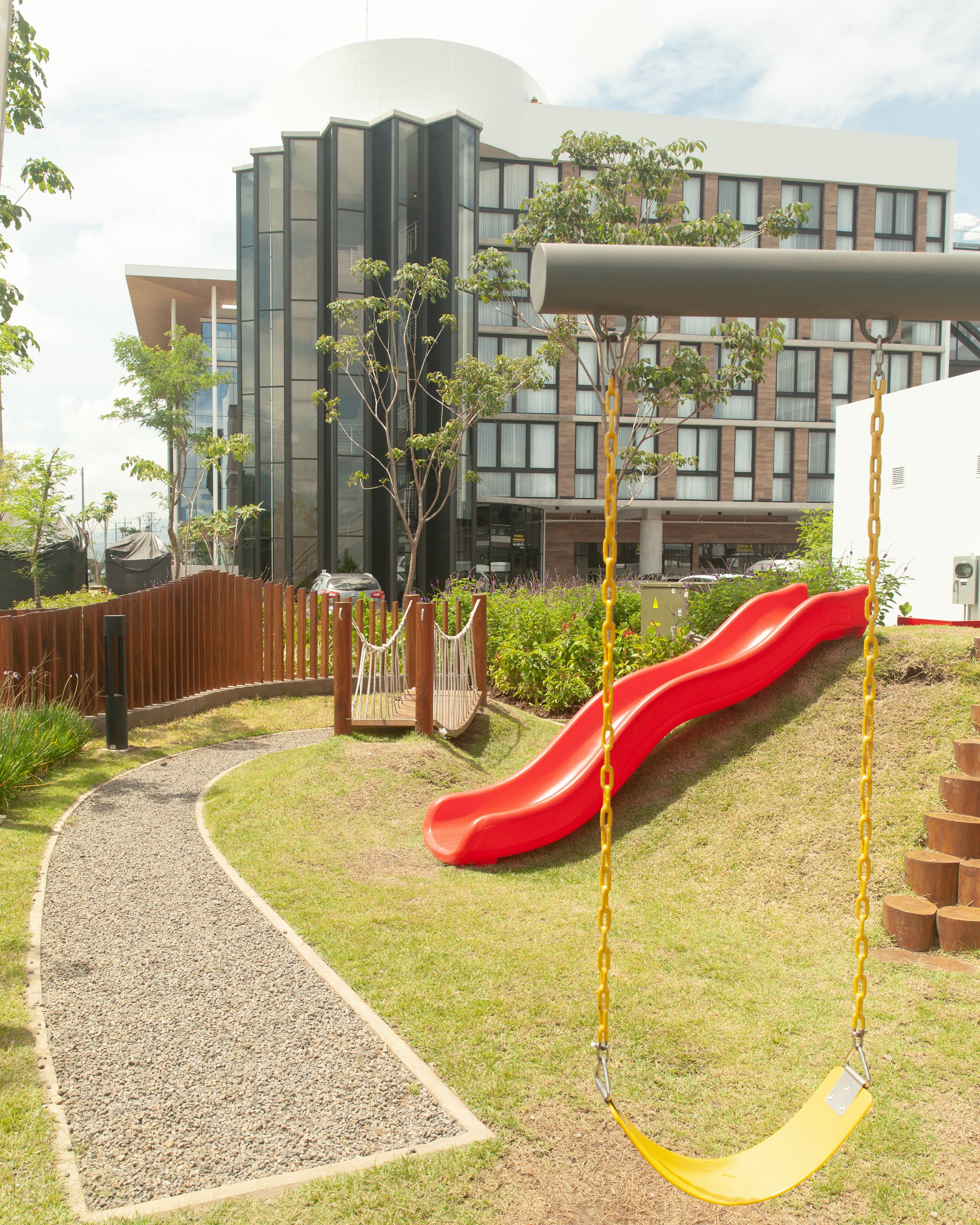 Un terrain de jeux avec un toboggan rouge et des balançoires, entouré d’un chemin de gravier et de verdure, avec un bâtiment moderne en arrière-plan.