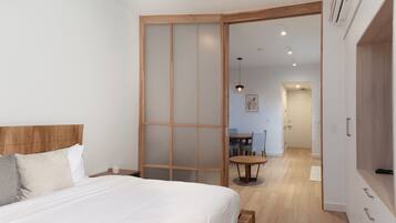 A modern bedroom with a wooden headboard, a bed with white linens, a small round table with a chair, and a sliding glass door leading to a dining area.