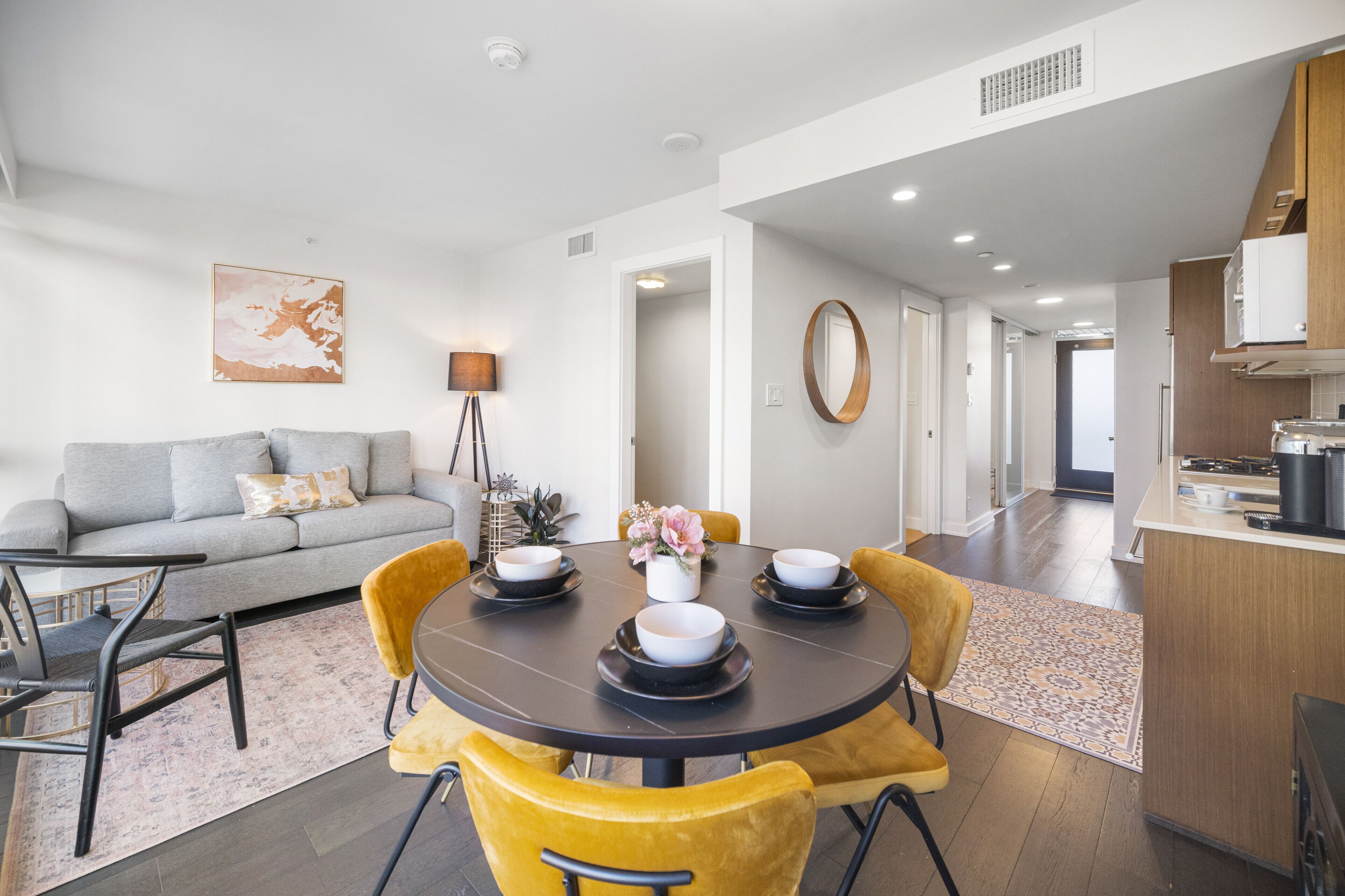 A modern living room with a grey sofa, yellow chairs, a round dining table with bowls, and a kitchen area with wooden cabinets.