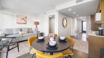A modern living room with a grey sofa, yellow chairs, a round dining table with bowls, and a kitchen area with wooden cabinets.