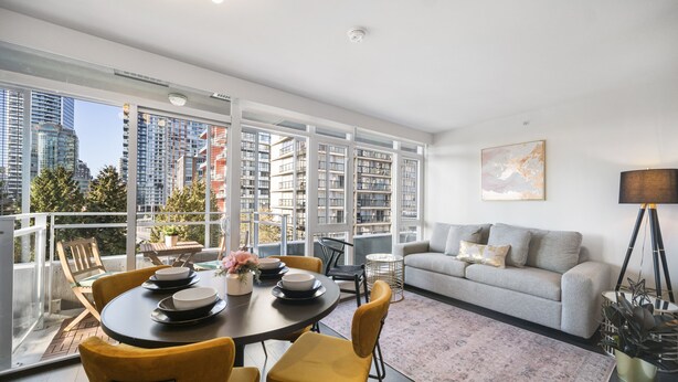 Ein modernes Wohnzimmer mit einem grauen Sofa, gelben Sesseln, einem Esstisch mit Tellern, einer Stehlampe und einem großen Fenster mit Blick auf die Stadt.