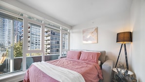 Ein Schlafzimmer mit großem Fenster, einem Bett mit rosa Bettwäsche, einem grauen Sofa, einer Stehlampe und einem gerahmten Kunstwerk an der Wand.