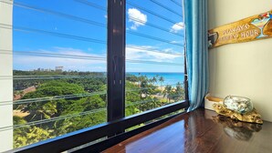 A room with a large window overlooking a beach and cityscape, a wooden sign with "ALWAYS HAPPY HOUR", and a glass bowl with seashells on a wooden table.