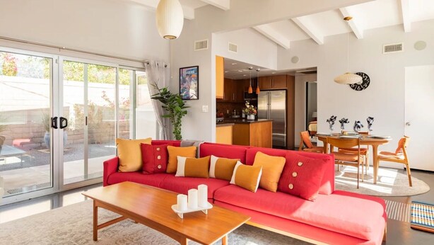 A modern living room with a red sectional sofa, a wooden coffee table, and a dining area with a wooden table and chairs. - Unique 2 Master Suite Pool/ Spa Home, Relaxing Yard with Mountain Views (Palm Springs)