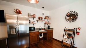 A kitchen with a stainless steel refrigerator, wooden cabinets, a sink, and a coffee machine. There are shelves with decorative items and a wall-mounted clock.