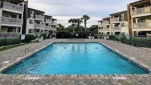A swimming pool surrounded by multi-story residential buildings with balconies, a clear sky, and a palm tree in the background. - stylish coastal condo with beautiful ocean views! (Myrtle Beach)
