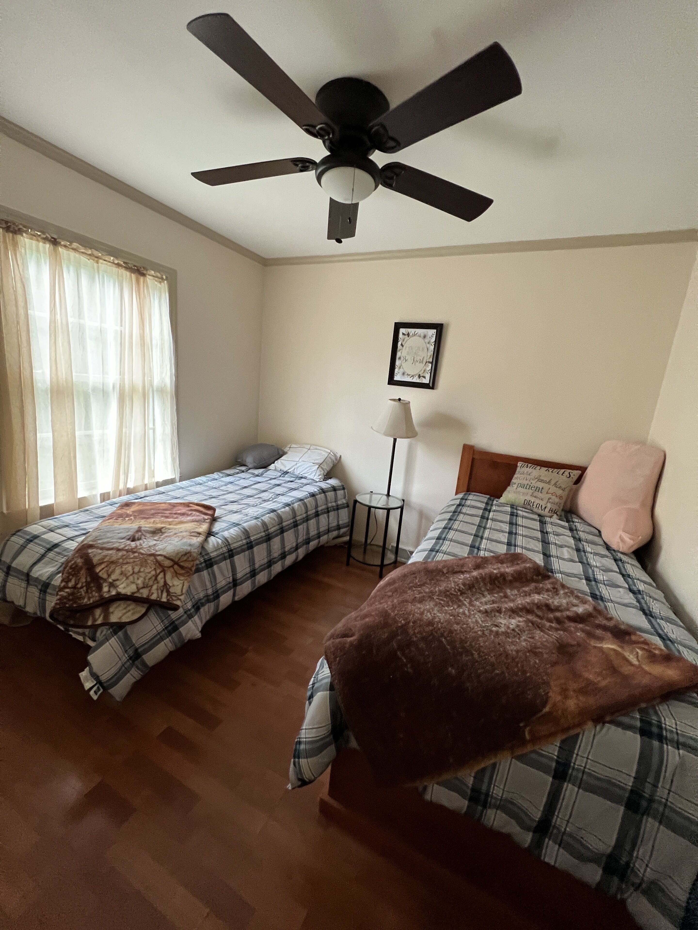 Ein Schlafzimmer mit zwei Betten, einem Deckenventilator, einem Nachttisch, einer Lampe und einem Fenster mit Vorhängen.