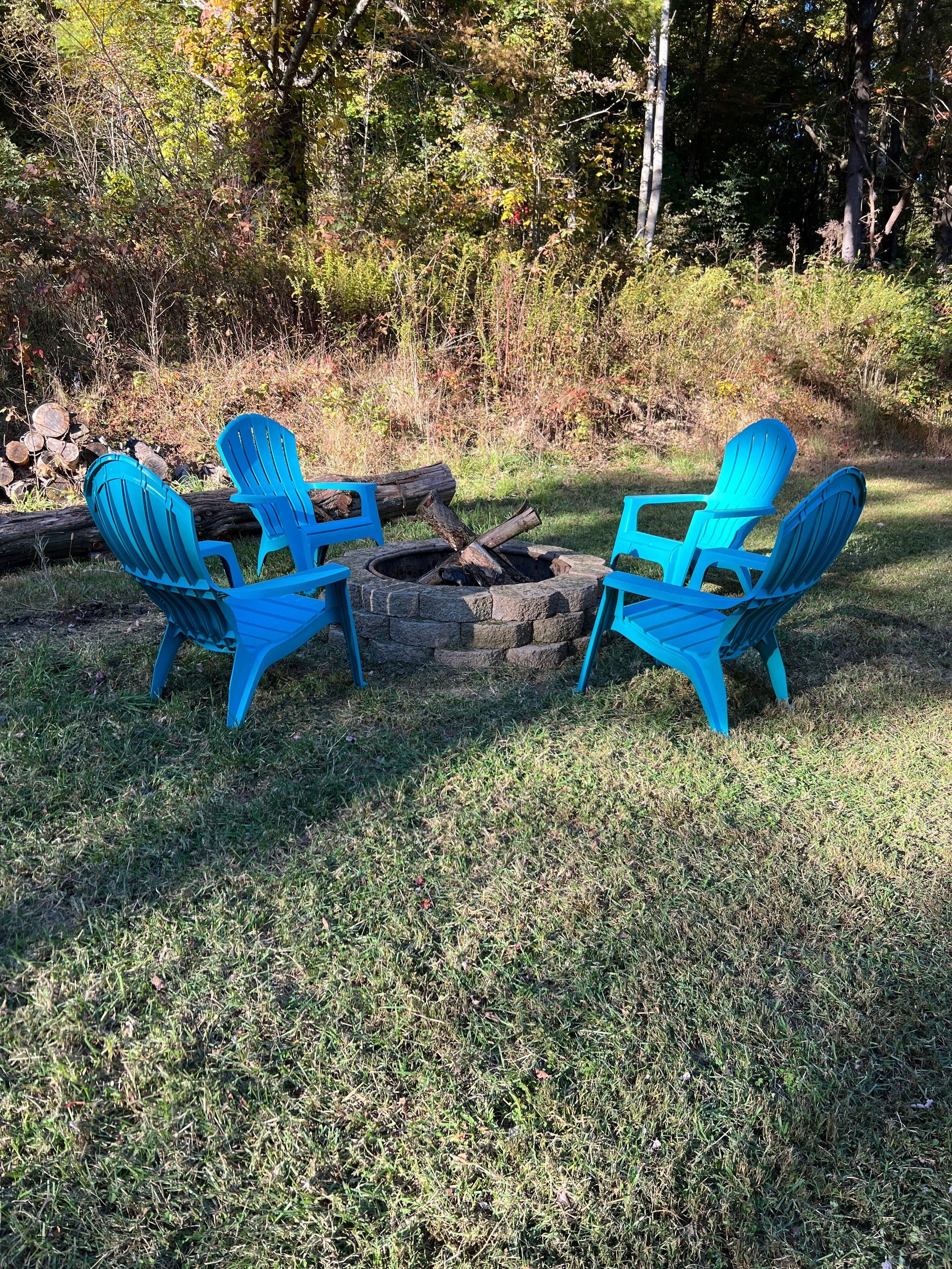 Two blue Adirondack chairs around a stone fire pit in a grassy outdoor area with trees in the background.