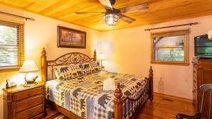 A bedroom with a wooden ceiling, a bed with a checkered bedspread, a ceiling fan, a bedside table with a lamp, a dresser, and a television on a stand.
